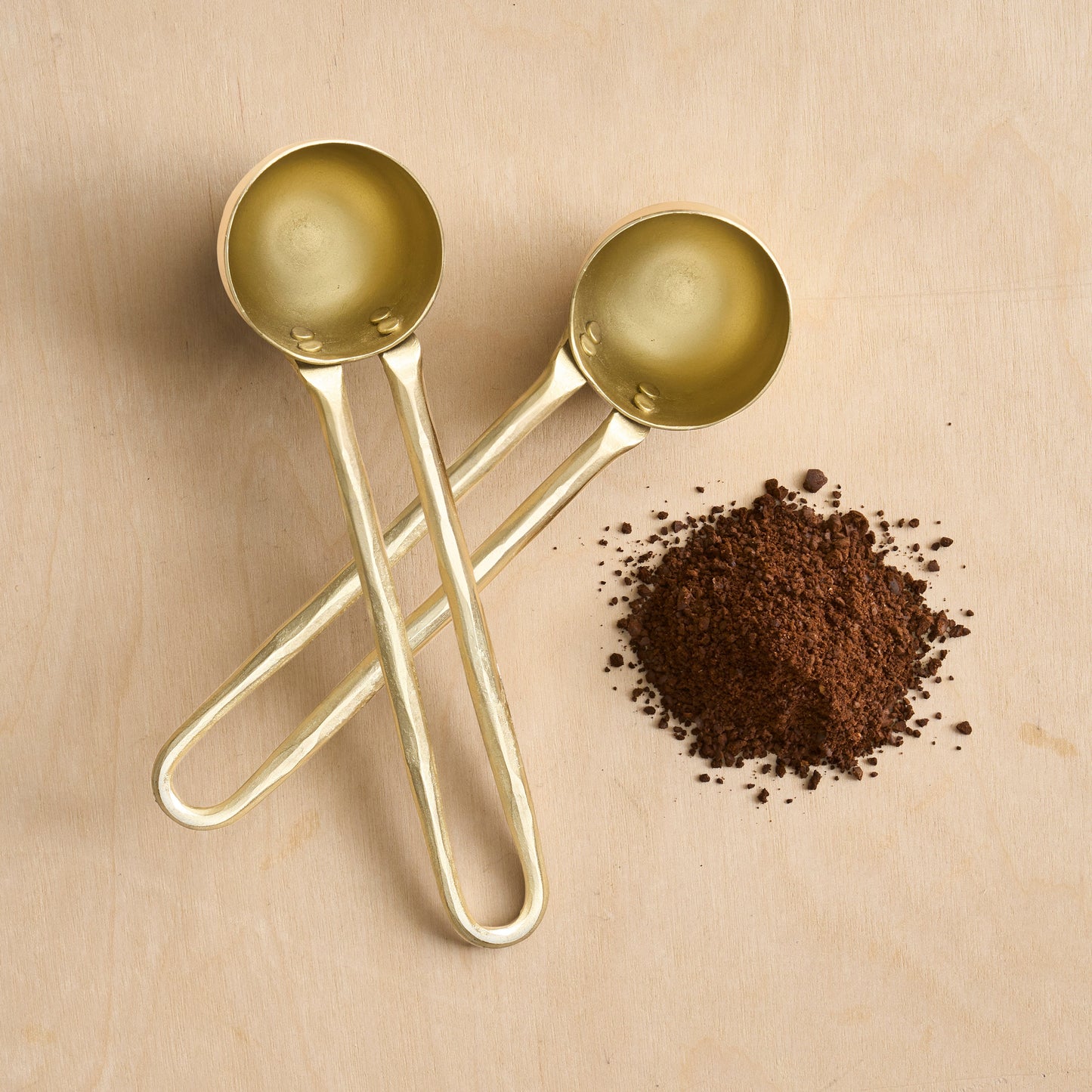 Two gold measuring spoons on a beige surface with coffee grounds.