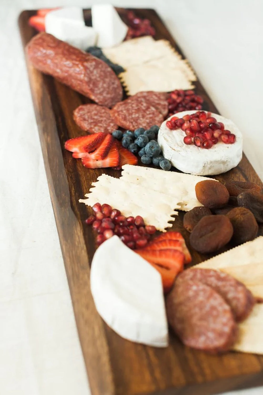 Wooden charcuterie board with various meats, cheeses, fruits, and nuts on a white background