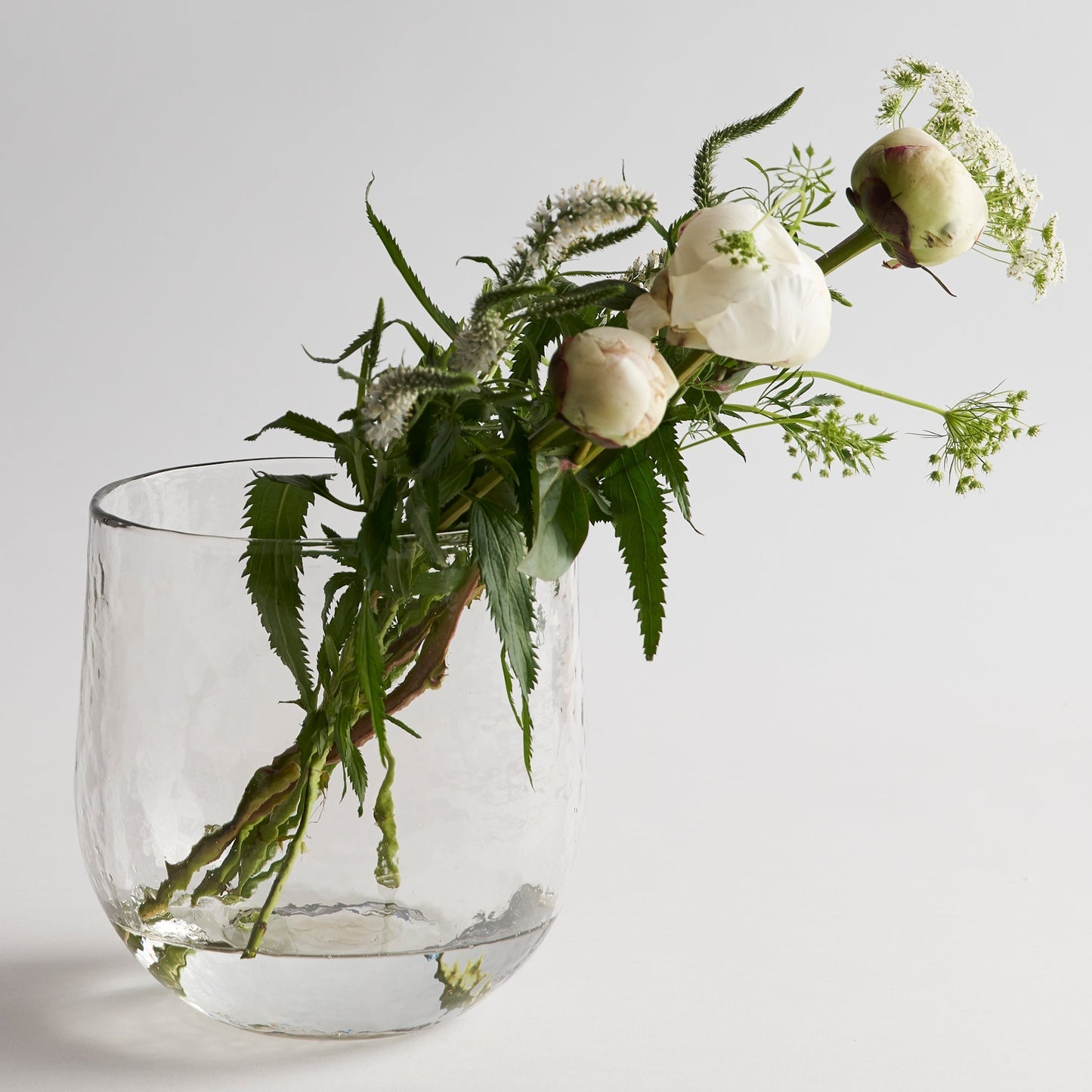 Clear glass vase with greenery and white flowers on a light gray background