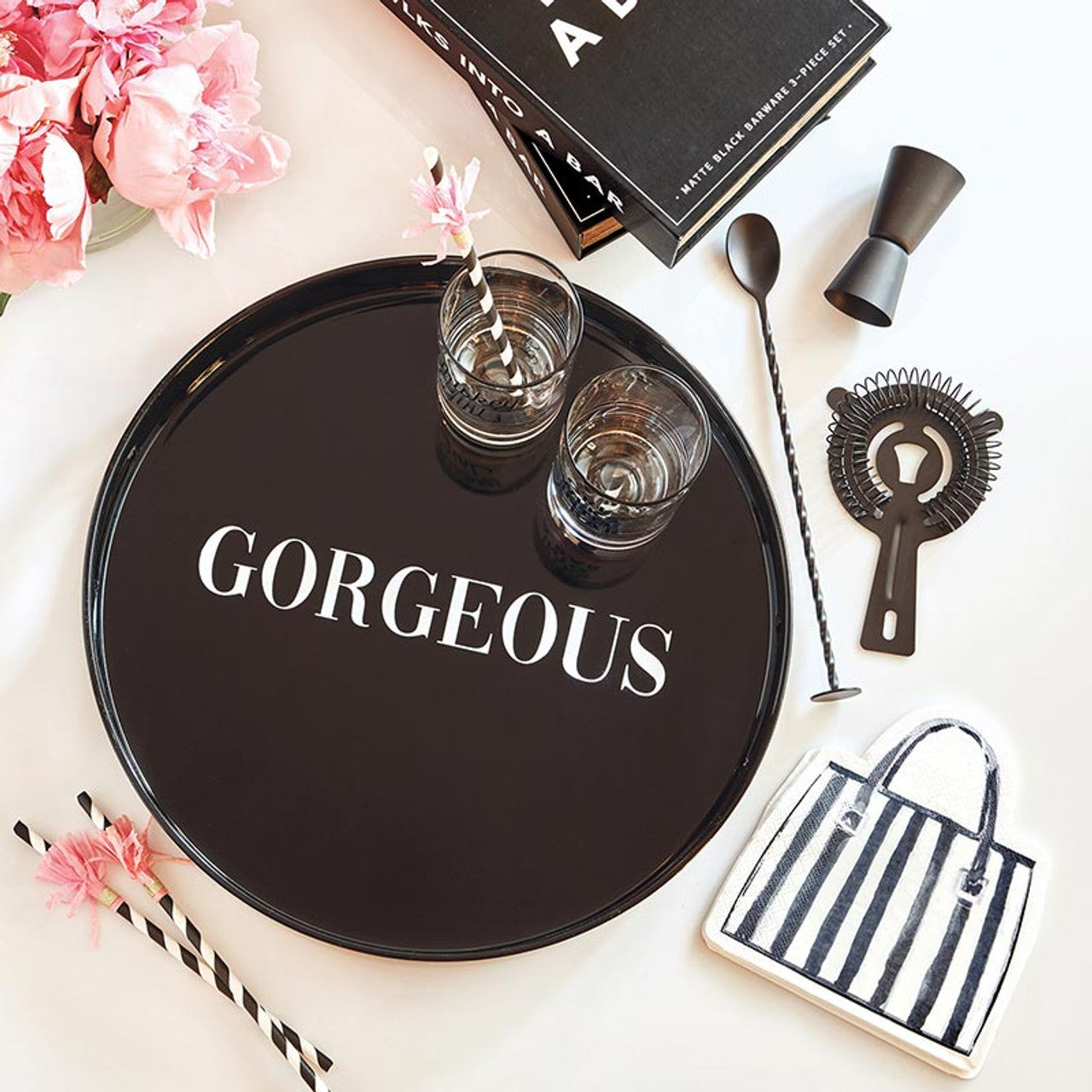 Black tray with 'GORGEOUS' text, glasses, flowers, and bar tools on a white surface