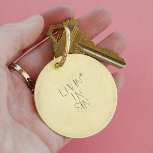 Gold keychain with 'Livin' in Sin' tag held by a hand against a pink background