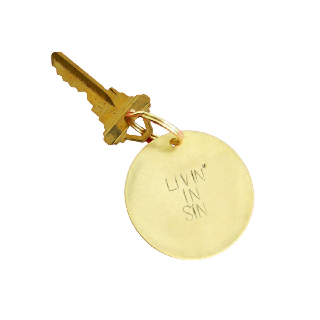 Gold key with a round tag engraved 'LIVIN' IN SIN' on a white background