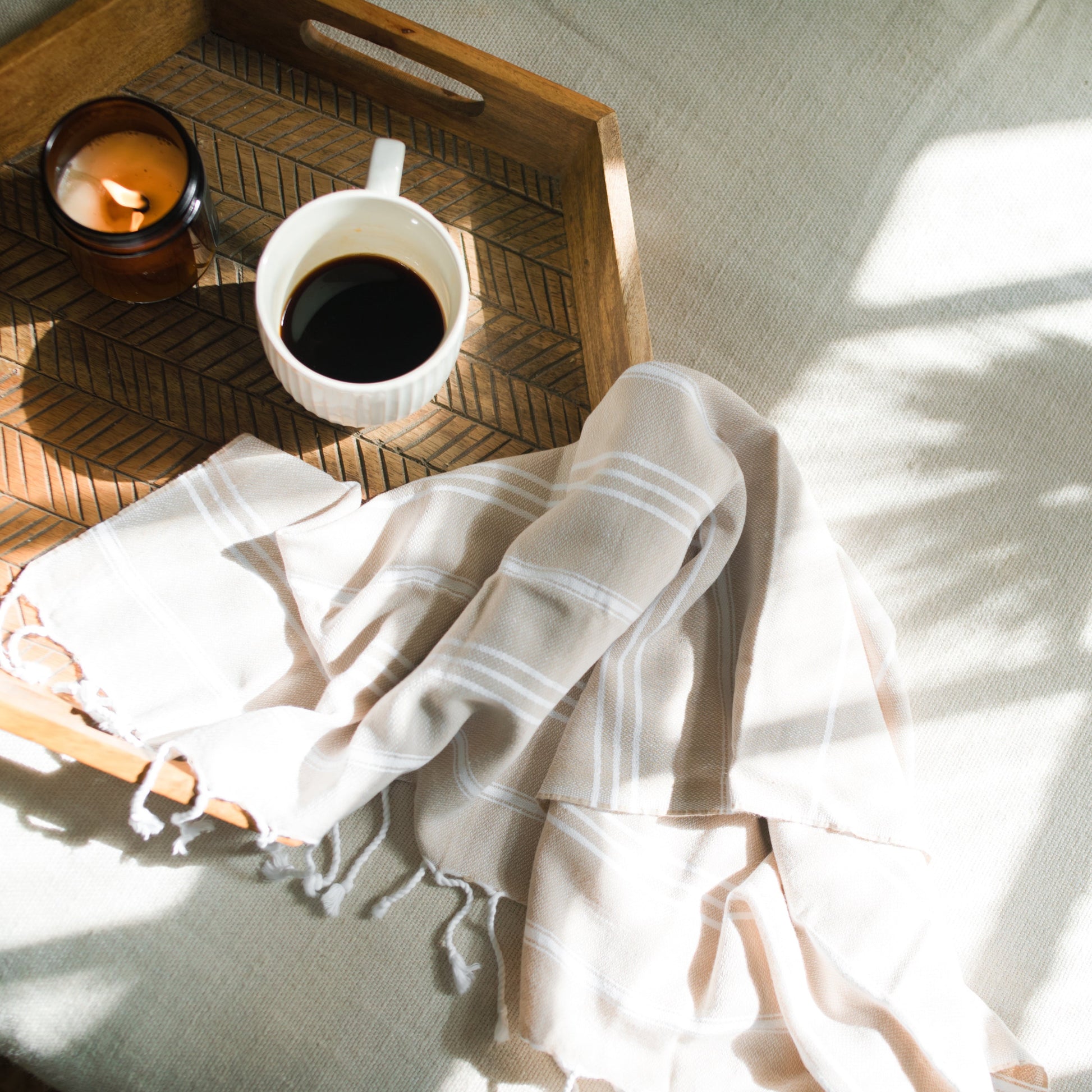 Wooden tray with a cup of coffee, candle, and white towel on a light surface.